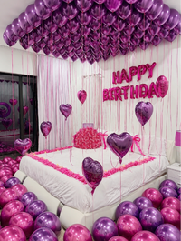 A decorated bedroom with a large bed covered in white bedding and pink rose petals, surrounded by many metallic purple and pink balloons. The wall behind the bed has pink foil balloons spelling “HAPPY BIRTHDAY,” with additional heart-shaped balloons and hanging pink ribbons.